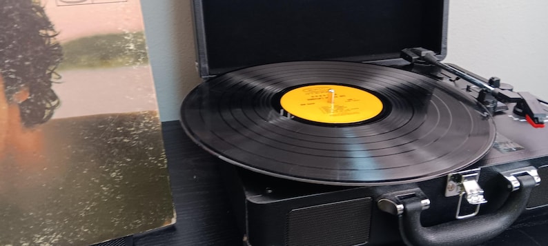 Pode incluir: Um disco de vinil preto tocando em um toca-discos preto vintage. O toca-discos est&aacute; aberto, com a agulha repousando sobre o vinil. Uma capa de &aacute;lbum &eacute; vis&iacute;vel ao fundo. O r&oacute;tulo do disco &eacute; amarelo.