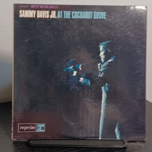 Puede incluir: Una portada de disco de vinilo cuadrada titulada Sammy Davis Jr. At The Cocoanut Grove. La portada presenta un fondo oscuro y texturizado con la silueta de un artista con traje. El logotipo de Reprise Records es visible en la esquina inferior izquierda.