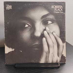 May include: A vintage vinyl record album cover featuring a black and white portrait of Roberta Flack. The album is titled "Chapter Two" and is displayed on a black stand. A black record player is visible in the background.