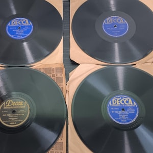 May include: Four black vinyl phonograph records, each housed in a tan paper sleeve. Three records feature blue Decca labels with white text, while one record features a gold Decca label with black text. The records are arranged in a two-by-two grid.