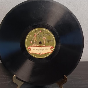 May include: A black circular vinyl record featuring a central label with green and tan accents. The label displays the text Official Boy Scout Record and Boy Scout Bugle Calls, along with an illustration of a scout figure in a uniform.
