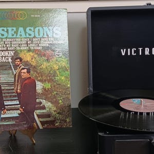 May include: Vintage vinyl record album cover for "The 4 Seasons" with the text "Lookin' Back" and a black Victrola record player. The album cover features a photo of the band members on a bridge. The record player is black with the word "Victrola" printed on the lid.