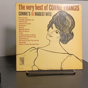 May include: A square vinyl record jacket titled The Very Best of Connie Francis, Connie's 15 Biggest Hits. The cover features a yellow background with a black line-art illustration of a woman and a tracklist of fifteen songs printed in black.