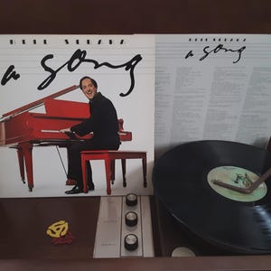 May include: Album cover for "A Song" by Barry Manilow, featuring a red piano and the artist. The album is open next to a record player with a vinyl record playing. The album cover has the title "a song" in cursive.