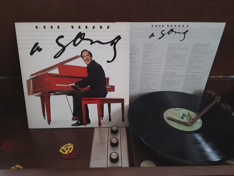 May include: Album cover for "A Song" by Barry Manilow, featuring a red piano and the artist. The album is open next to a record player with a vinyl record playing. The album cover has the title "a song" in cursive.