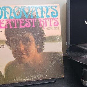 Pode incluir: Um &aacute;lbum de vinil vintage intitulado "Donovan's Greatest Hits" com um retrato de Donovan. Ao lado, um toca-discos Victrola preto com um disco girando. A capa do &aacute;lbum tem texto turquesa e rosa.