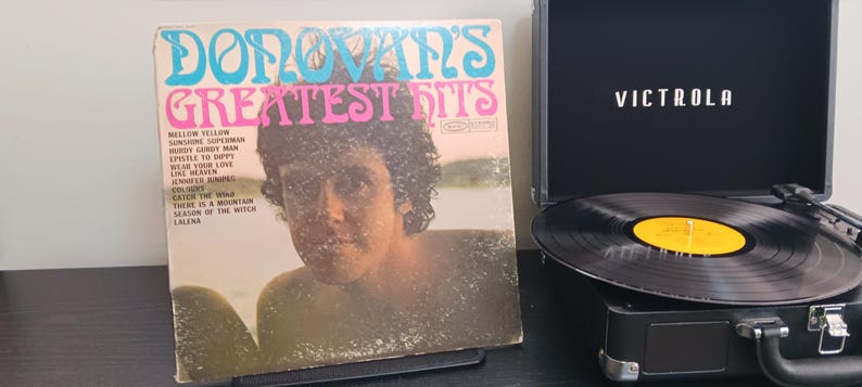 Pode incluir: Um &aacute;lbum de vinil vintage intitulado "Donovan's Greatest Hits" com um retrato de Donovan. Ao lado, um toca-discos Victrola preto com um disco girando. A capa do &aacute;lbum tem texto turquesa e rosa.