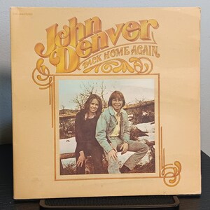 May include: A square vinyl record album cover featuring a tan background with the text John Denver Back Home Again in stylized gold lettering. A central rectangular photograph shows two people sitting outdoors on a wooden fence in a rural setting.