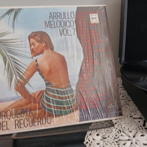 May include: A vintage vinyl record album cover featuring a woman in a swimsuit on a beach with palm trees. The album is titled "Arrullo Melodico Vol. 7" and features the text "Orquesta del Recuerdo".
