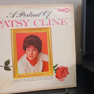 May include: Vintage vinyl record album titled "A Portrait of Patsy Cline." The album cover features a portrait of Patsy Cline in a gold frame, with a red rose and the word "DECCA" on the top right corner. A black Victrola record player is in the background.