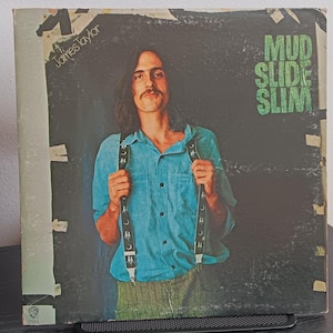 May include: Vintage vinyl record album cover featuring James Taylor. The album cover has a dark background with the words "Mud Slide Slim" in green. Taylor is wearing a blue shirt and suspenders.