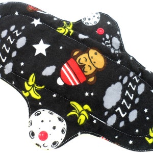 Op de afbeelding: Een zwarte herbruikbare stoffen menstruatiepad met een kleurrijke print van apen, bananen, sterren en manen. De pad heeft een rode knop aan de zijkant.