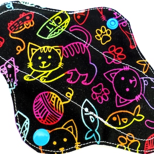 Puede incluir: Una compresa de tela reutilizable negra con un estampado colorido de gatos y peces. La compresa tiene dos botones a presión azules.