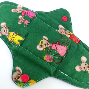 Peut inclure: Un protège-slip en tissu réutilisable de couleur verte avec une bordure blanche cousue et un motif répétitif de souris dans différentes tenues. Le protège-slip a deux boutons-pression rouges sur les côtés.