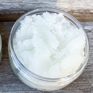 May include: A silver jar with a white label that reads "Mediterranean Sea Salt Scrub - Eucalyptus Mint -" and lists ingredients. The jar is filled with a white, grainy scrub. The jar is sitting on a wooden surface.