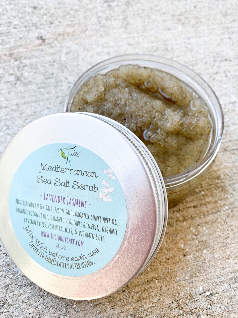 May include: A silver tin with a white label that reads "Jule Mediterranean Sea Salt Scrub - Lavender Jasmine - Mediterranean Sea Salt, Epsom Salt, Organic Sunflower Oil, Organic Coconut Oil, Organic Vegetable Glycerin, Organic Lavender Buds, Essential Oils, & Vitamin E Oil. www.tulebodycare.com 8 oz Mix Well before each use. Cover Lid Immediately After Using."
