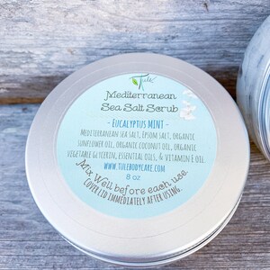 May include: A silver tin with a blue label that reads "Mediterranean Sea Salt Scrub - Eucalyptus Mint - Mediterranean Sea Salt, Epsom Salt, Organic Sunflower Oil, Organic Coconut Oil, Organic Vegetable Glycerin, Essential Oils, & Vitamin E Oil. www.tulebodycare.com 8 oz Mix Well before each use. Cover lid immediately after using."