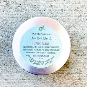 May include: Silver metal jar with a white label that reads "Jule Mediterranean Sea Salt Scrub - Lavender Jasmine - Mediterranean Sea Salt, Epsom Salt, Organic Sunflower Oil, Organic Coconut Oil, Organic Vegetable Glycerin, Organic Lavender Buds, Essential Oils, & Vitamin E Oil. www.tulebodycare.com 8 oz Mix Well before each use. Cover lid immediately after using."