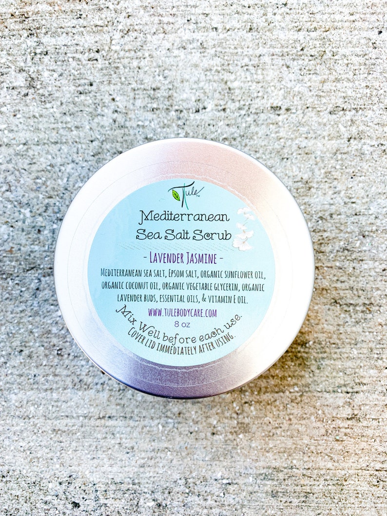 May include: Silver metal jar with a white label that reads "Jule Mediterranean Sea Salt Scrub - Lavender Jasmine - Mediterranean Sea Salt, Epsom Salt, Organic Sunflower Oil, Organic Coconut Oil, Organic Vegetable Glycerin, Organic Lavender Buds, Essential Oils, & Vitamin E Oil. www.tulebodycare.com 8 oz Mix Well before each use. Cover lid immediately after using."