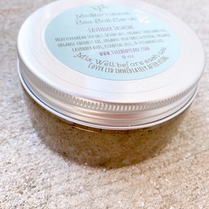 May include: A silver jar with a light blue label that reads "Mediterranean Sea Salt Scrub - Lavender Jasmine". The label also includes the ingredients, website, and instructions to "Mix Well before each use. Cover lid immediately after using." The jar is 8 oz and contains a brown, grainy scrub.