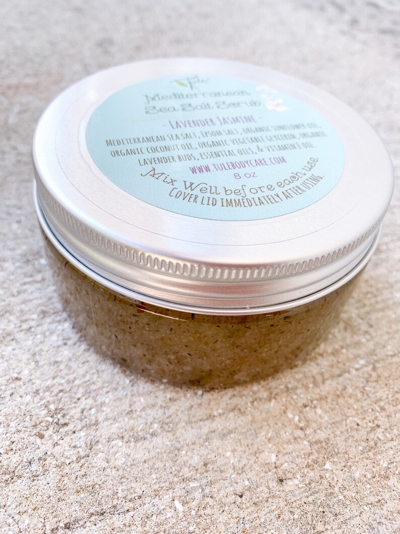 May include: A silver jar with a light blue label that reads "Mediterranean Sea Salt Scrub - Lavender Jasmine". The label also includes the ingredients, website, and instructions to "Mix Well before each use. Cover lid immediately after using." The jar is 8 oz and contains a brown, grainy scrub.