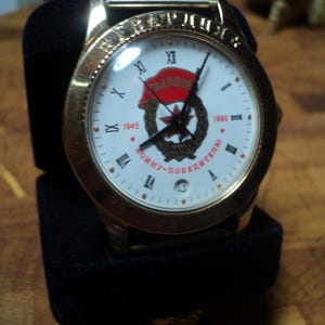 May include: Gold-toned wristwatch with a white face featuring Roman numerals and a red star emblem. The watch is displayed in a black velvet box. The watch face includes the text "1945" and "1995".