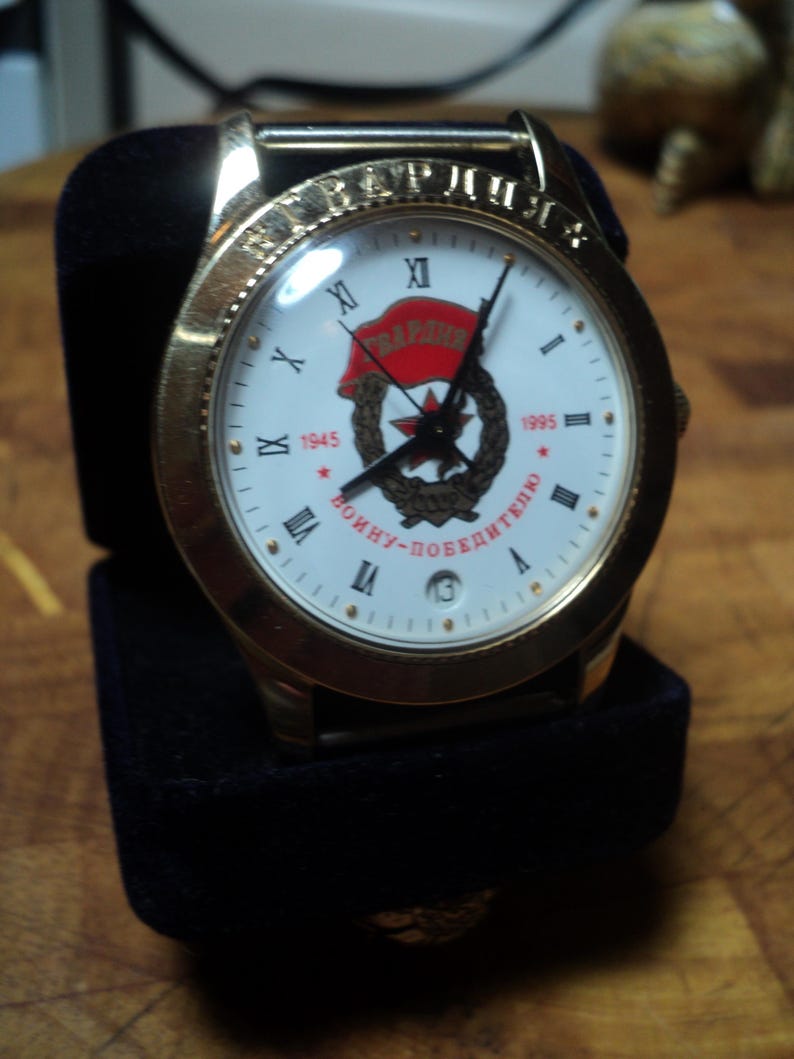 May include: Gold-toned wristwatch with a white face featuring Roman numerals and a red star emblem. The watch is displayed in a black velvet box. The watch face includes the text "1945" and "1995".