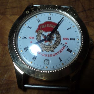 May include: A gold-toned wristwatch with a white face featuring Roman numerals and the text "ГВАРДИЯ". The watch face includes a red star and the years 1945 and 1995. The watch has a date display at the 6 o'clock position.