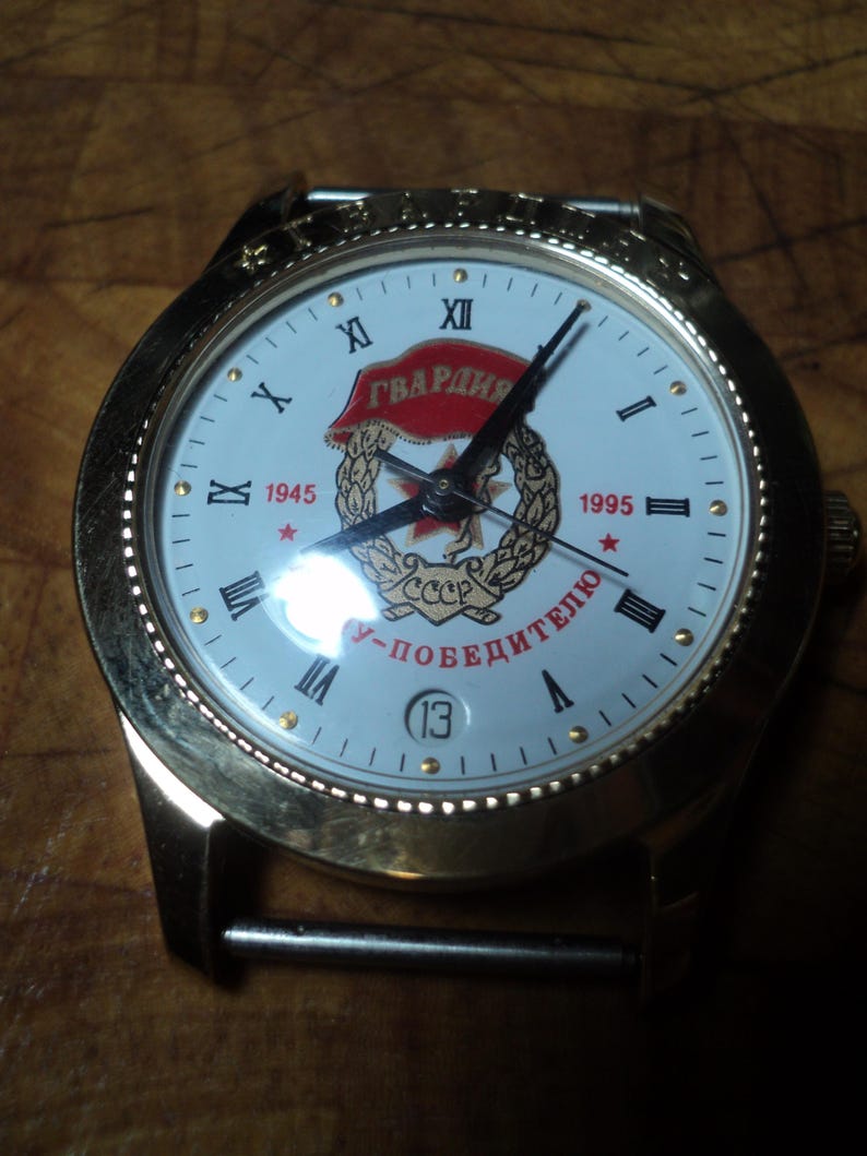 May include: A gold-toned wristwatch with a white face featuring Roman numerals and the text "ГВАРДИЯ". The watch face includes a red star and the years 1945 and 1995. The watch has a date display at the 6 o'clock position.