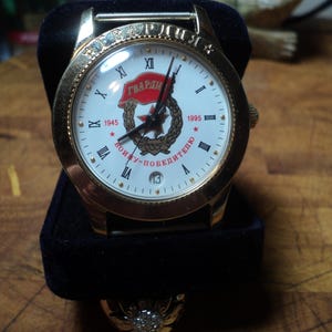 May include: Gold-toned wristwatch with a white face featuring Roman numerals and the text "ГВАРДИЯ". The watch has a black band and a date display at the 3 o'clock position. The watch is presented on a black velvet stand.