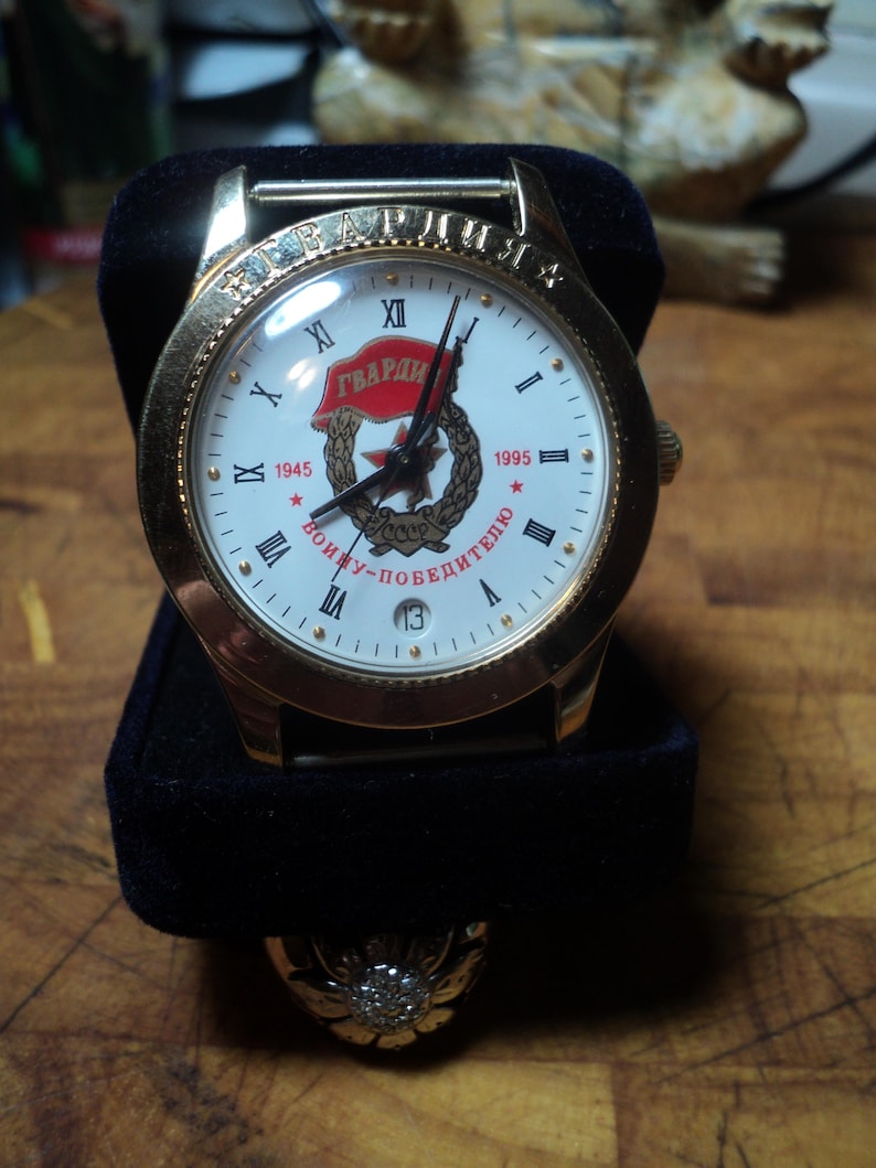 May include: Gold-toned wristwatch with a white face featuring Roman numerals and the text "ГВАРДИЯ". The watch has a black band and a date display at the 3 o'clock position. The watch is presented on a black velvet stand.