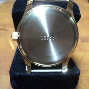 May include: A gold-toned wristwatch with a silver-colored back and the number "169380" engraved on it. The watch is displayed on a dark blue velvet surface, showcasing its polished metal finish and classic design.