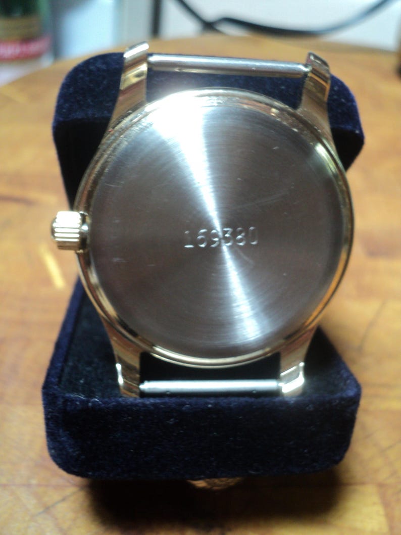 May include: A gold-toned wristwatch with a silver-colored back and the number "169380" engraved on it. The watch is displayed on a dark blue velvet surface, showcasing its polished metal finish and classic design.