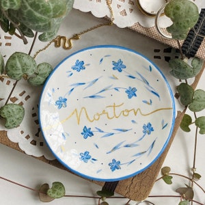 May include: A small, white ceramic dish with a blue rim, decorated with blue floral designs and the gold word "Norton". The dish is surrounded by greenery and a gold snake necklace.