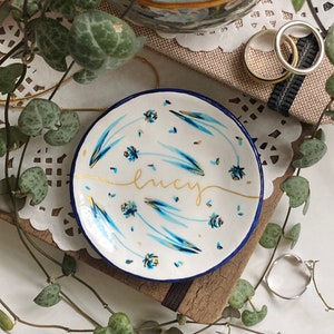 May include: A white ceramic ring dish with a blue and gold floral design. The dish has the name "Lucy" written in gold script on the front.