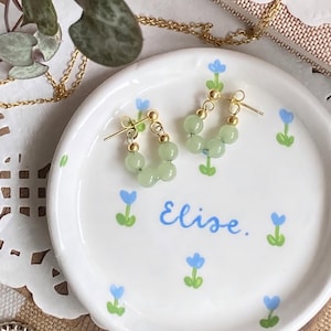 May include: A pair of gold hoop earrings with light green beads. The earrings are on a white ceramic dish with blue flowers and the name "Elise" written in cursive.