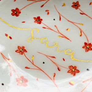 May include: A white ceramic dish with a gold inscription of the name "Sara" and a floral pattern of red flowers and branches.