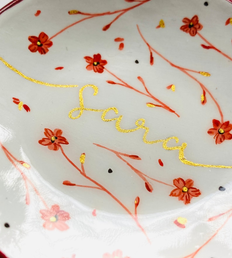 May include: A white ceramic dish with a gold inscription of the name "Sara" and a floral pattern of red flowers and branches.