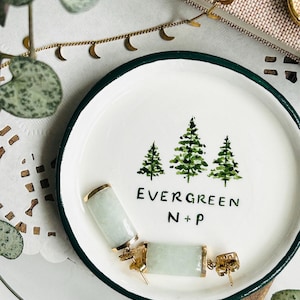 Puede incluir: Un plato de cerámica blanco con borde verde y una pintura a la acuarela de tres árboles de hoja perenne. El plato tiene el texto "EVERGREEN N+P". Dos pendientes rectangulares de jade con detalles dorados están en el plato.