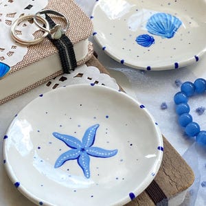 Op de afbeelding: Twee kleine keramische schalen met blauwe en witte ontwerpen. Een schaal heeft een blauwe zeester met witte accenten, terwijl de andere een blauwe schelp met witte accenten heeft. Beide schalen hebben een blauw gestippeld patroon rond de rand.