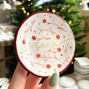 May include: A white ceramic trinket dish with a red rim. The dish is decorated with a floral pattern in red and gold. The name "Sara" is written in gold script on the dish.