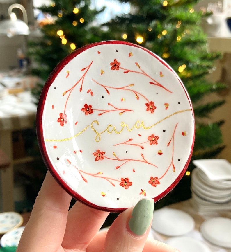 May include: A white ceramic trinket dish with a red rim. The dish is decorated with a floral pattern in red and gold. The name "Sara" is written in gold script on the dish.
