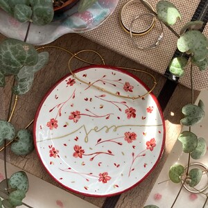 May include: A white ceramic trinket dish with a red rim and a floral design in shades of red and gold. The word "Jess" is written in gold script on the dish.