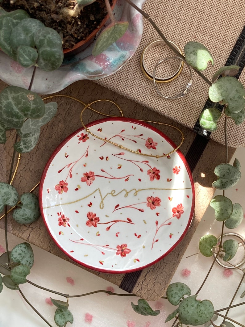 May include: A white ceramic trinket dish with a red rim and a floral design in shades of red and gold. The word "Jess" is written in gold script on the dish.