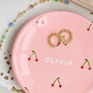 Puede incluir: Un plato de cerámica rosa con un motivo de cerezas y el nombre "Olivia" escrito en blanco. Dos aros de oro están en el plato.