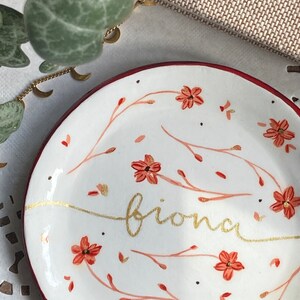 May include: A white ceramic trinket dish with a red rim. The dish is decorated with a floral pattern in shades of red and gold. The word "brionna" is written in gold script across the center of the dish.