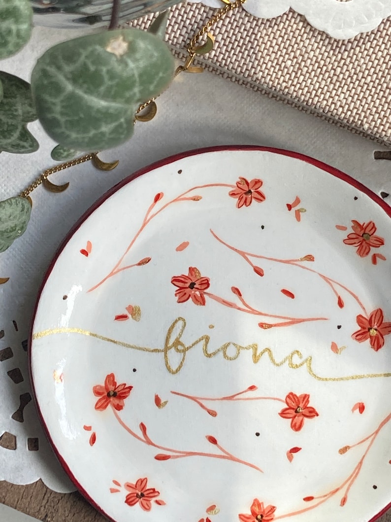May include: A white ceramic trinket dish with a red rim. The dish is decorated with a floral pattern in shades of red and gold. The word "brionna" is written in gold script across the center of the dish.
