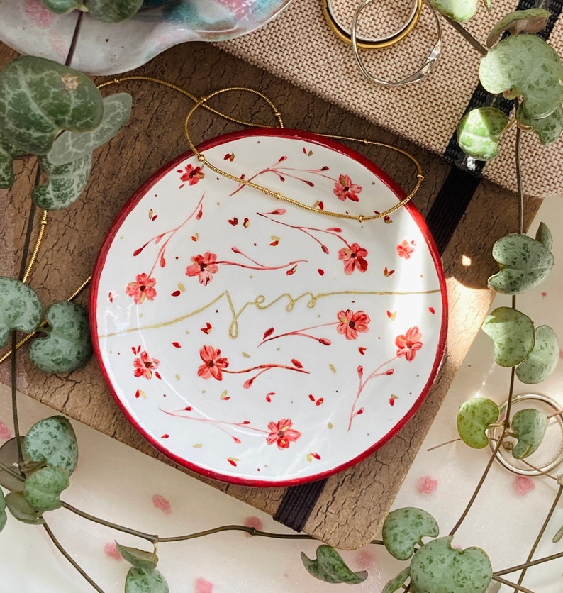May include: A white ceramic ring dish with a red rim and a floral design. The dish has the word "Jess" written in gold script.