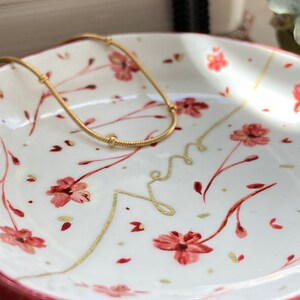 May include: A white ceramic dish with a pink rim and a floral design. The dish has a gold chain necklace resting on it.