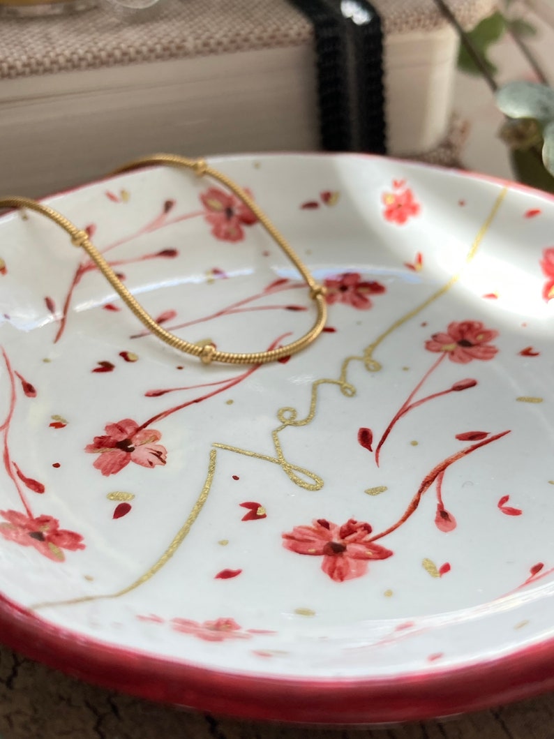 May include: A white ceramic dish with a pink rim and a floral design. The dish has a gold chain necklace resting on it.